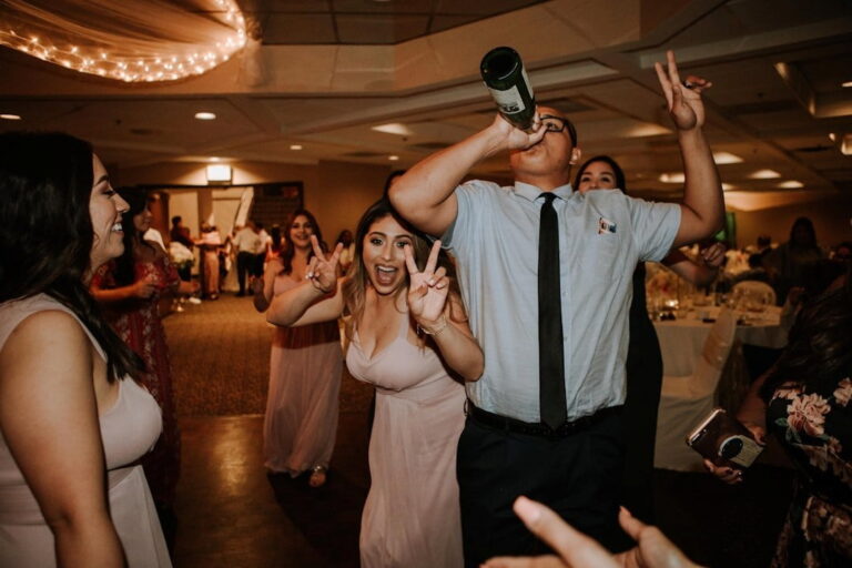 Man drinking out of wine bottle next to woman who appears to be a bridesmaid, facing the camera and giving a peace sign at wedding venues St. Louis
