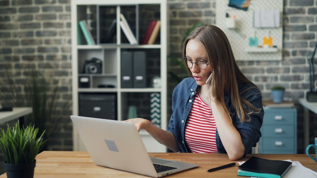 Woman with glasses sitting at a desk, staring at a laptop, and talking on a cell phone