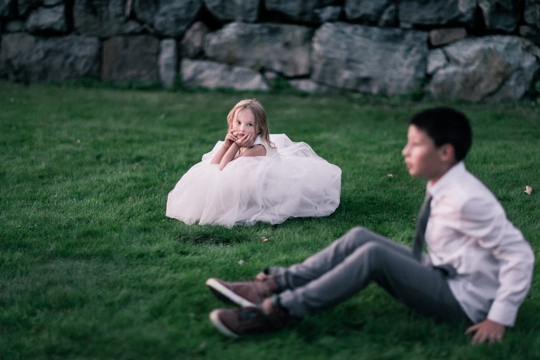 Young girl in a while flower girl dress sitting in a grassy area staring at a boy also sitting